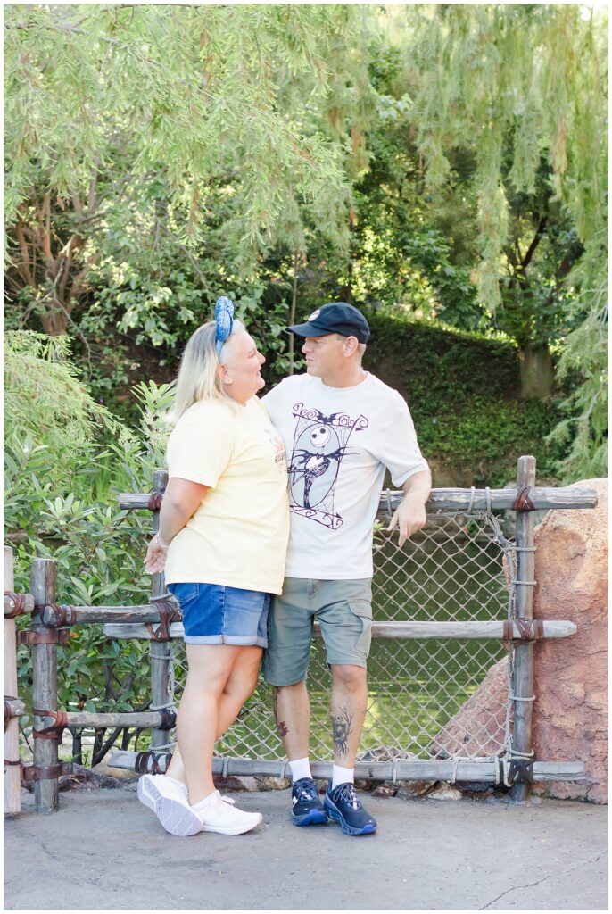 A couple stands close together, looking into each other's eyes, with lush green trees in the background. The woman is wearing a yellow shirt and denim shorts, while the man wears a white "Nightmare Before Christmas" shirt and green shorts. They are standing near a wooden fence.