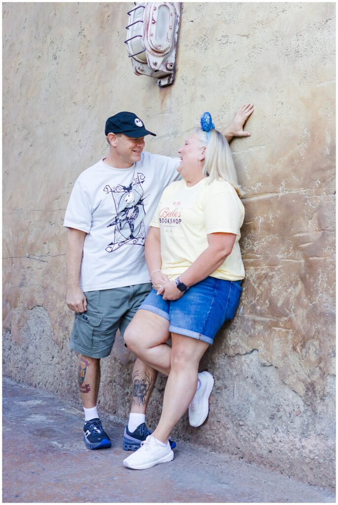 The couple leans against a textured wall, smiling at each other. The woman, in a yellow shirt and denim shorts, rests her hand on the wall, while the man, in a white "Nightmare Before Christmas" shirt and green shorts, casually leans next to her.