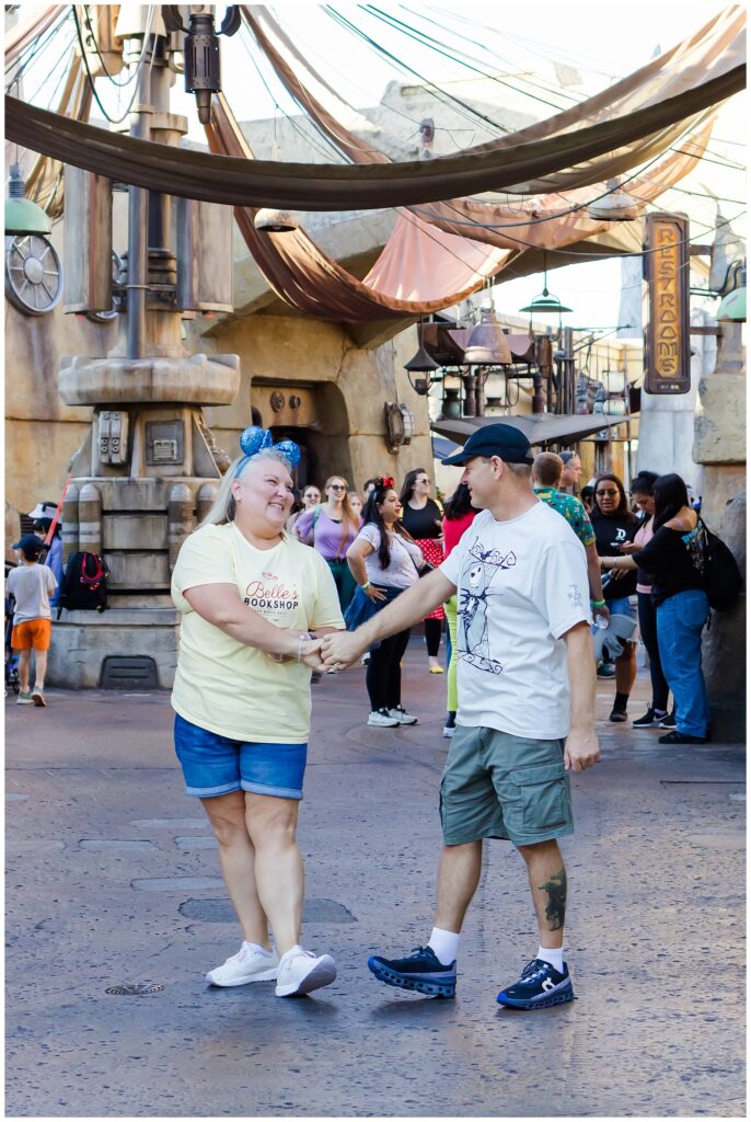 The couple holds hands and dances in a bustling theme park setting. The woman wears a yellow shirt and denim shorts, and the man is in a white "Nightmare Before Christmas" shirt and green shorts. They are surrounded by other park visitors.