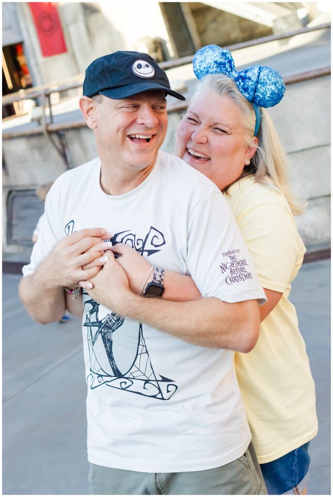 The couple embraces playfully, laughing together. The woman, in a yellow shirt and blue Minnie Mouse ears, hugs the man from behind. He wears a white "Nightmare Before Christmas" shirt and a black cap.