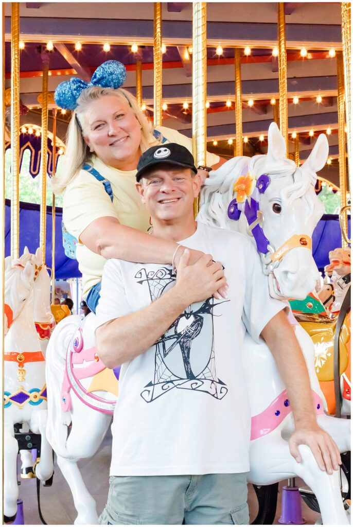 The couple poses on a carousel, with the woman seated behind the man, smiling brightly. The man stands in front of her, holding a carousel pole. Both are dressed in casual attire, with the woman in a yellow shirt and blue Minnie Mouse ears, and the man in a white "Nightmare Before Christmas" shirt.