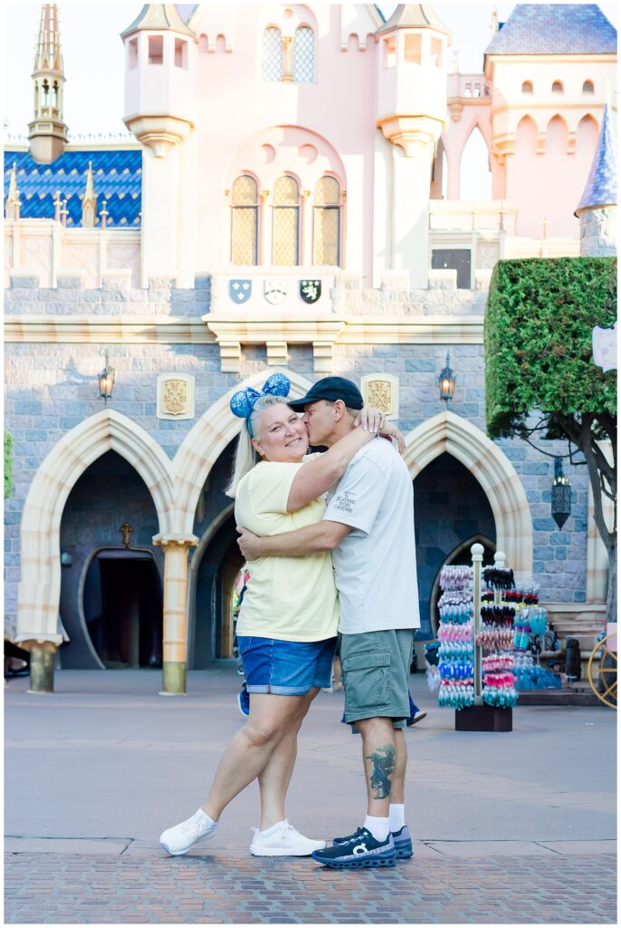 The couple stands in front of a castle, embracing each other. The woman wears a yellow shirt and blue Minnie Mouse ears, while the man wears a white "Nightmare Before Christmas" shirt and green shorts. The man kisses the woman on the cheek.