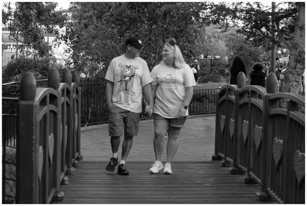 The couple walks hand-in-hand across a wooden bridge, smiling at each other. The image is in black and white. The woman is in a yellow shirt and denim shorts, and the man is in a white "Nightmare Before Christmas" shirt and green shorts.