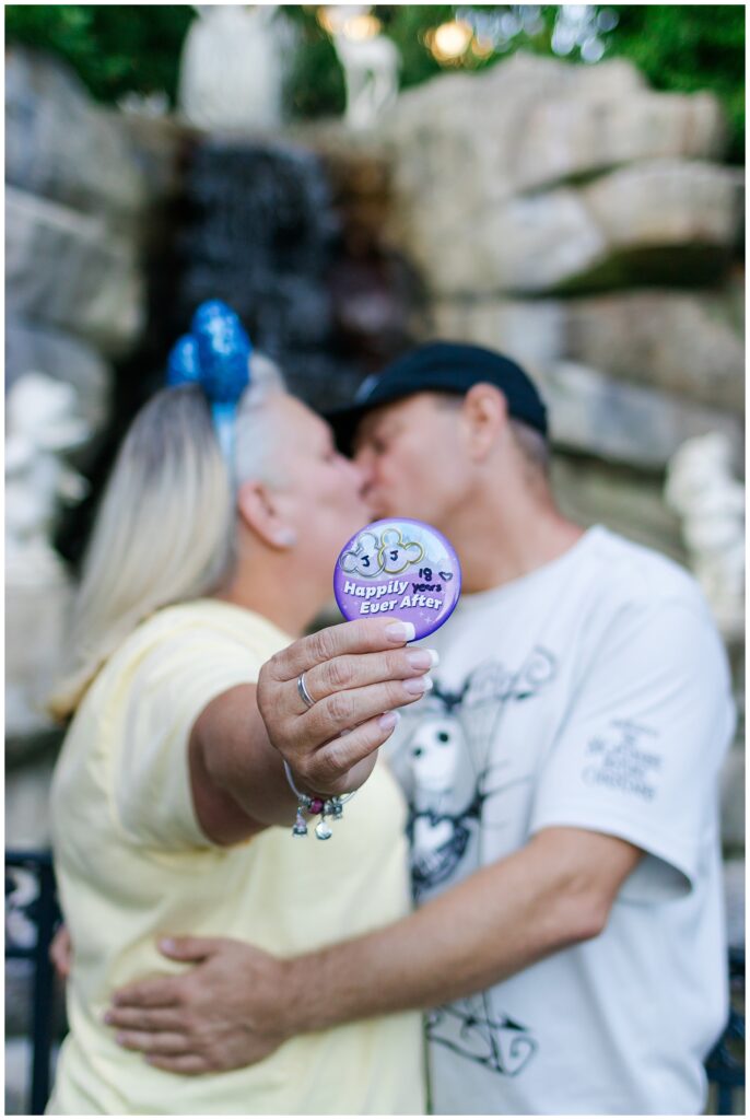 The couple kisses in front of a waterfall, with the woman holding a "Happily Ever After" button towards the camera. The woman is in a yellow shirt and blue Minnie Mouse ears, and the man is in a white "Nightmare Before Christmas" shirt and black cap.