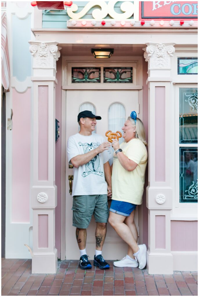 The couple stands in front of a pastel-colored building, sharing a Mickey Mouse-shaped pretzel. The woman wears a yellow shirt and denim shorts, while the man is in a white "Nightmare Before Christmas" shirt and green shorts. They hold hands and smile at each other.