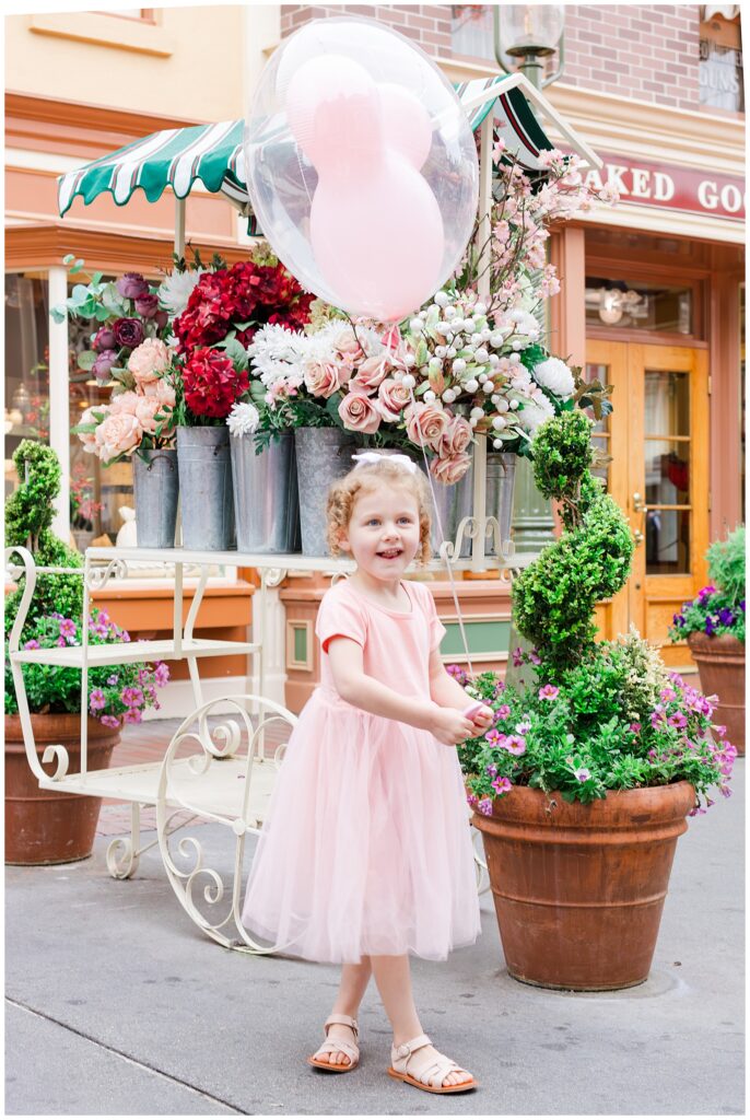 A young girl in a pink dress holding a clear balloon with pink balloons inside, standing in front of a cart adorned with colorful flowers.