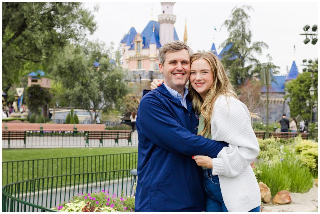 A smiling couple embracing, with the Disneyland castle in the background, surrounded by greenery and flowers.