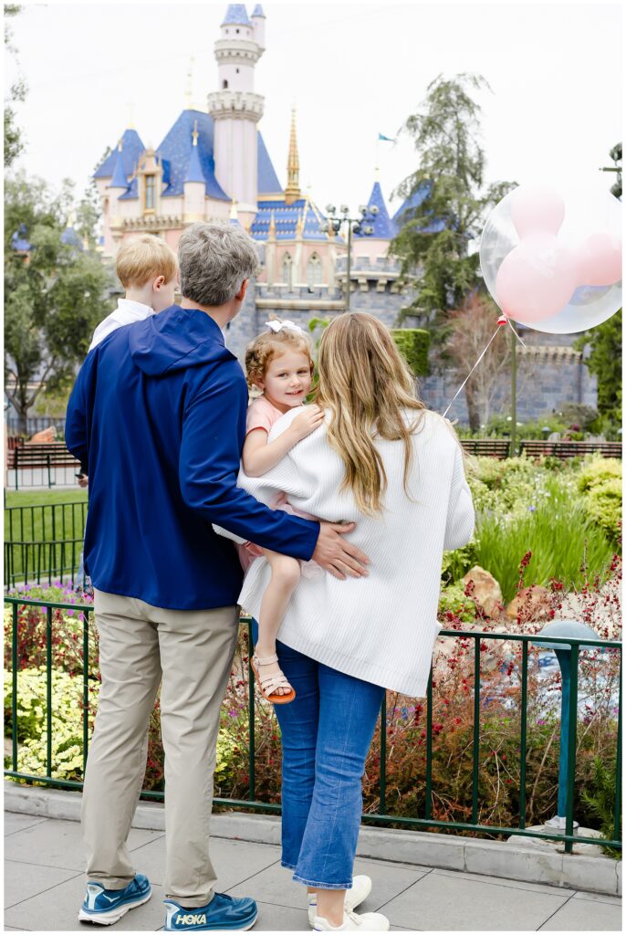 A family of four, with parents holding their young children, facing the Disneyland castle. The mother holds the daughter and the father holds the son.