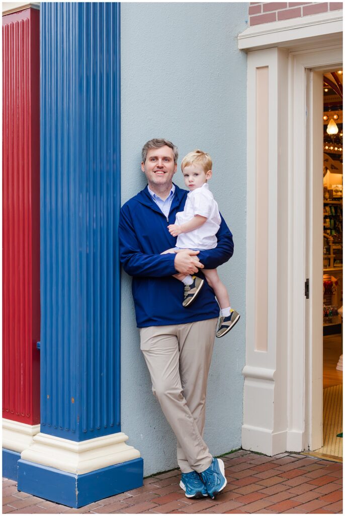 A father holding his young son while leaning against a building with red, blue, and gray columns.