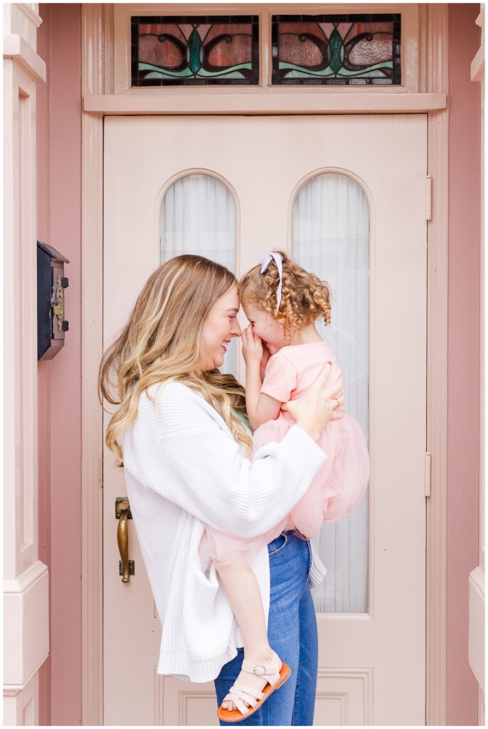 A mother and daughter sharing a tender moment, with the mother lifting the daughter who is covering her face shyly, standing in front of a pink door.