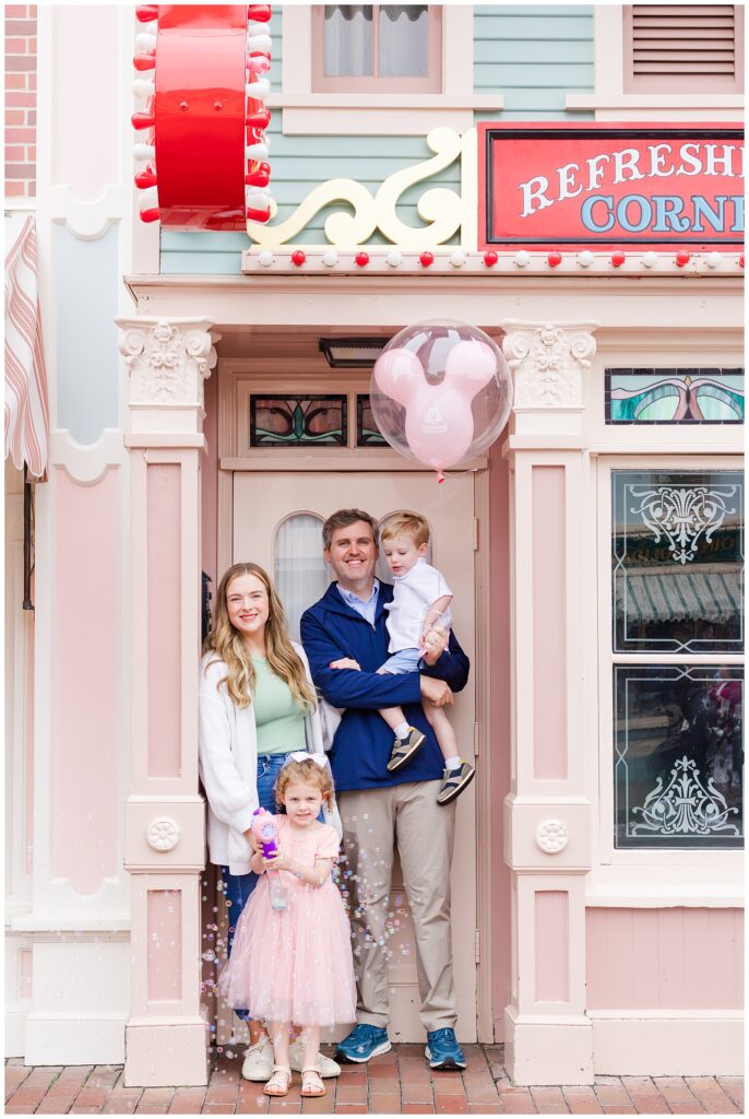 A family of four standing in front of a pastel-colored building with a sign reading "Refreshment Corner." The father holds the son and the daughter stands in front holding a bubble wand.