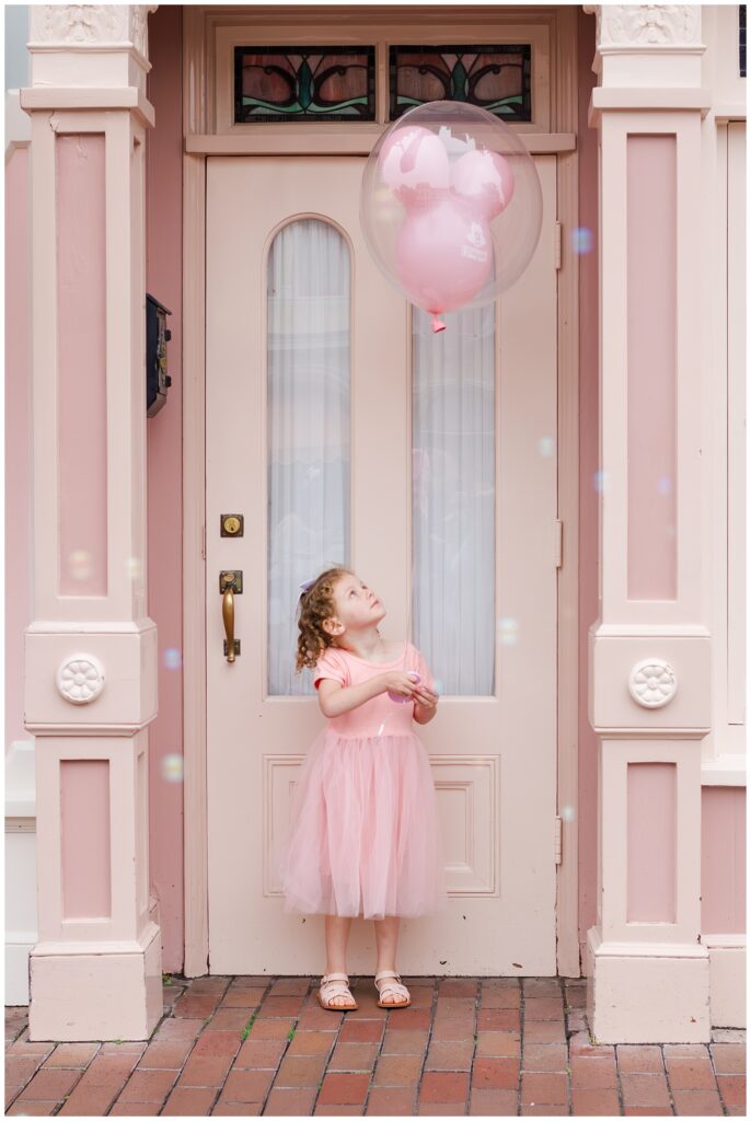 A young girl in a pink dress looking up at a clear balloon with pink balloons inside, standing in front of a pink door.