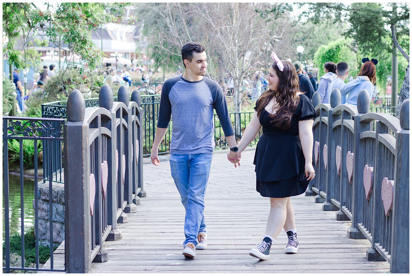 A couple holding hands and walking across a wooden bridge with heart-shaped decorations on the rails. The woman is wearing a black dress and pink Minnie Mouse ears, while the man is wearing a grey baseball shirt and blue jeans. They are smiling at each other with trees and other people visible in the background.