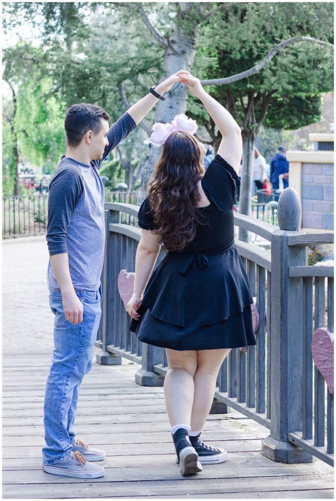 A couple on a wooden bridge, with the man twirling the woman. The woman is wearing a black dress and pink Minnie Mouse ears, while the man is in a grey baseball shirt and blue jeans. They are surrounded by trees and greenery, with other visitors in the background.