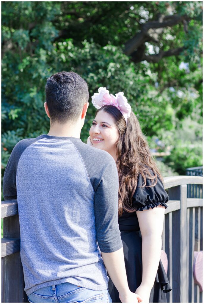 A couple standing close together on a wooden bridge. The woman, wearing a black dress and pink Minnie Mouse ears, is looking lovingly at the man, who is in a grey baseball shirt and blue jeans. Trees and greenery form the backdrop.