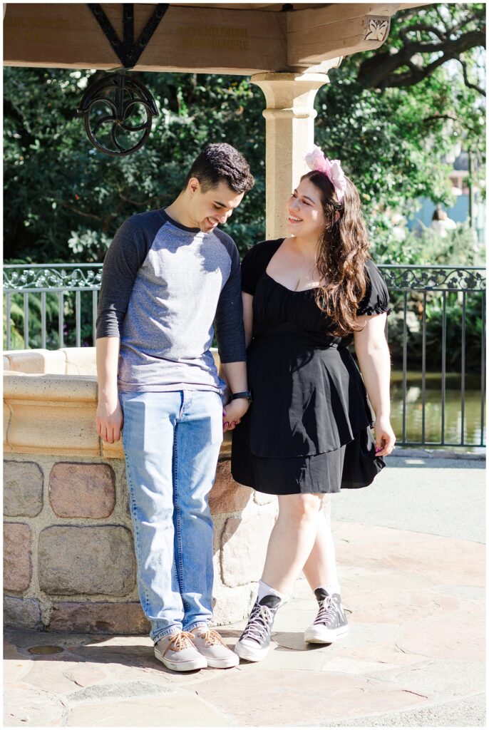 A couple standing in front of a stone structure, holding hands. The woman is in a black dress and pink Minnie Mouse ears, and the man is wearing a grey baseball shirt and blue jeans. They are both smiling and looking at each other.