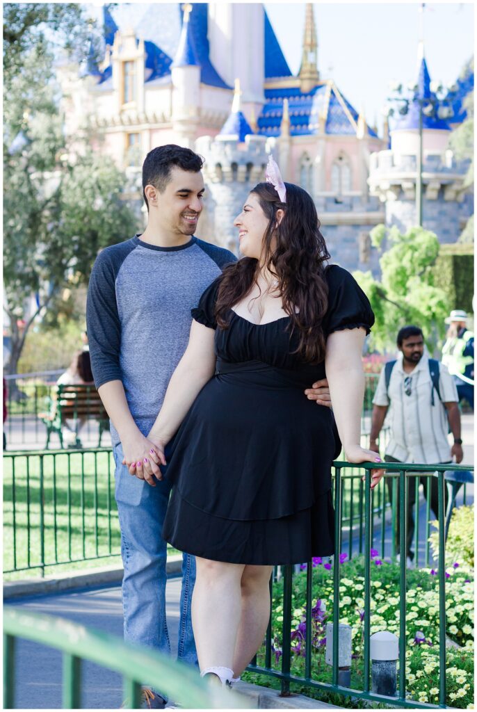 A couple standing in front of a green fence with a fairy tale castle in the background. The woman is wearing a black dress and pink Minnie Mouse ears, while the man is in a grey baseball shirt and blue jeans. They are holding hands and smiling at each other.