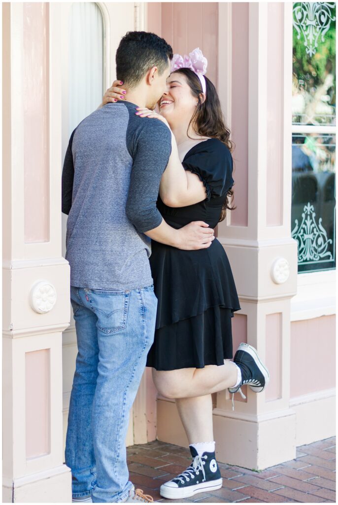 A couple standing close together and embracing in front of a pink building with decorative windows. The woman is wearing a black dress and pink Minnie Mouse ears, and the man is in a grey baseball shirt and blue jeans. They are both smiling and leaning in for a kiss.