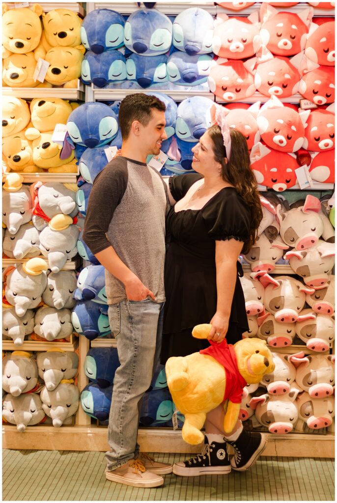 A couple standing in front of a wall filled with colorful stuffed animals, including Winnie the Pooh, Stitch, and Pua from Moana. The woman, holding a large Winnie the Pooh plush, is wearing a black dress and pink Minnie Mouse ears, while the man is in a grey baseball shirt and blue jeans. They are smiling at each other.