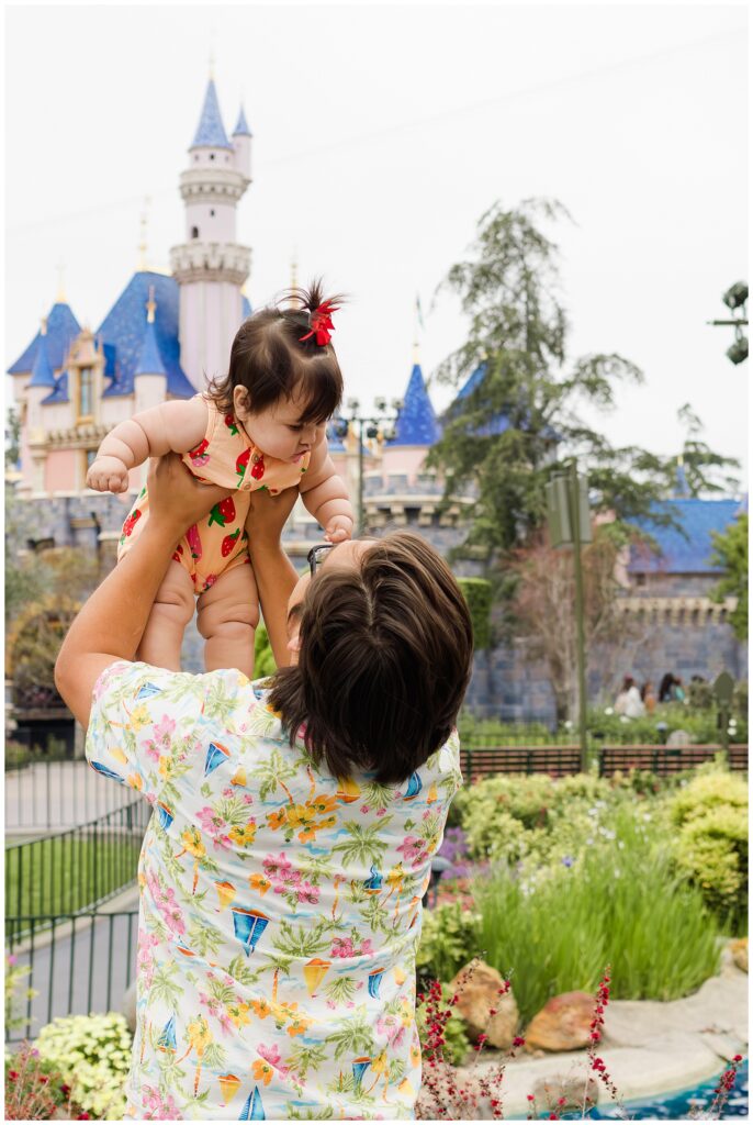 A father lifts his baby girl into the air in front of Sleeping Beauty Castle at Disneyland. The baby is wearing a strawberry-patterned romper and a red bow in her hair, while the father sports a colorful floral shirt.