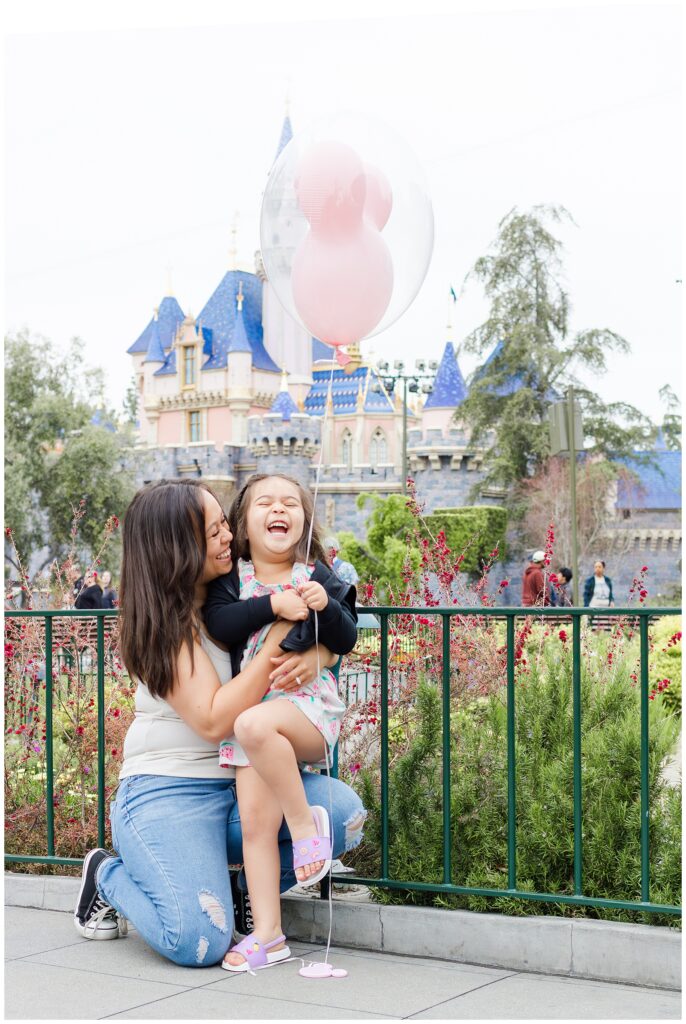 A mother kneels and hugs her laughing daughter, who is holding a Mickey Mouse balloon, in front of Sleeping Beauty Castle at Disneyland. The mother is dressed casually in a tank top and jeans, and the daughter is in a watermelon-patterned dress with a black jacket.