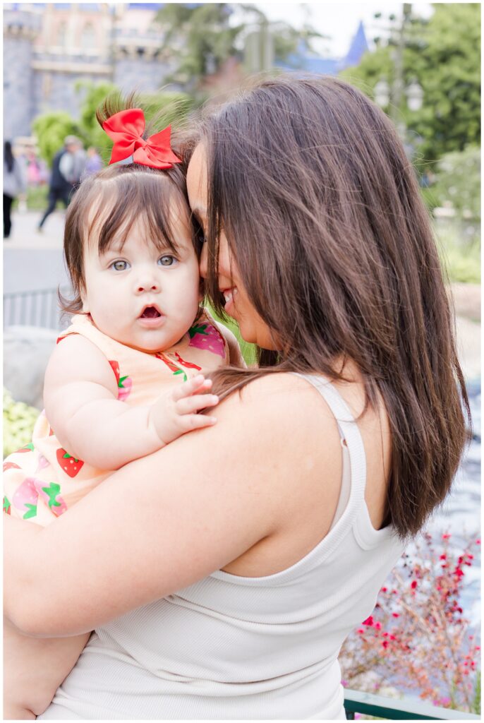 A mother holds her baby close to her chest with Sleeping Beauty Castle in the background at Disneyland. The baby, dressed in a strawberry-patterned romper with a red bow, looks curiously at the camera, while the mother smiles warmly.