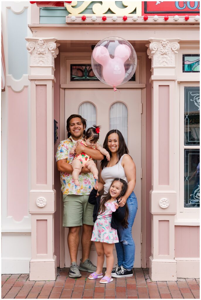 A family of four stands in front of a pink storefront at Disneyland. The father holds the baby, who wears a strawberry-patterned romper, and the mother hugs their older daughter, who holds a Mickey Mouse balloon. Everyone is smiling and happy.