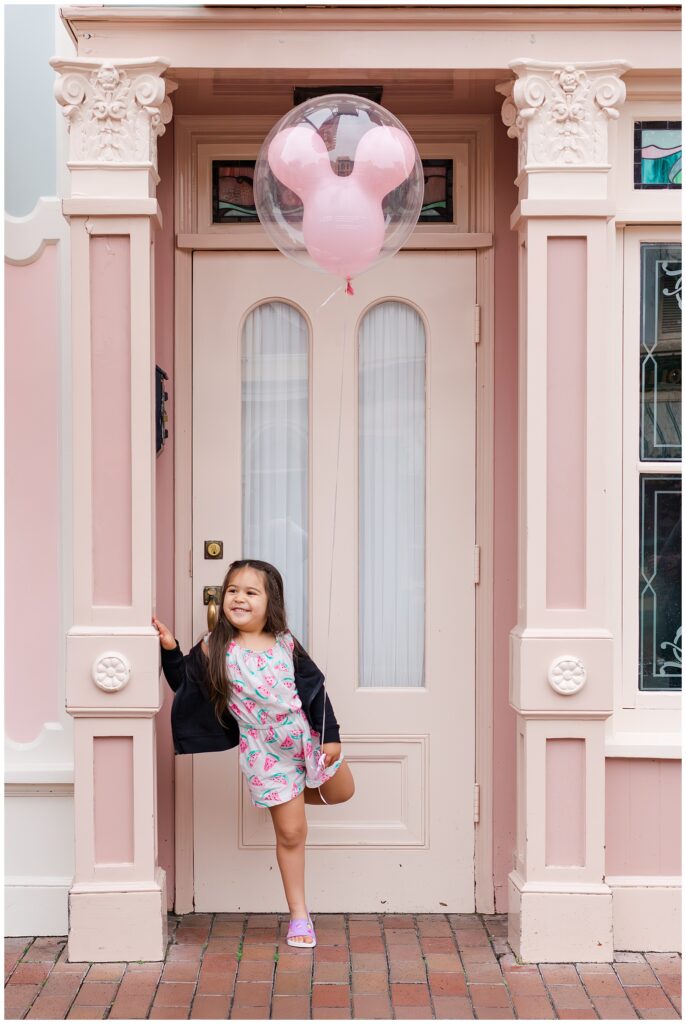 A young girl in a watermelon-patterned dress poses in front of a pink storefront at Disneyland, holding a Mickey Mouse balloon. She has a big smile on her face and is leaning against the door frame.