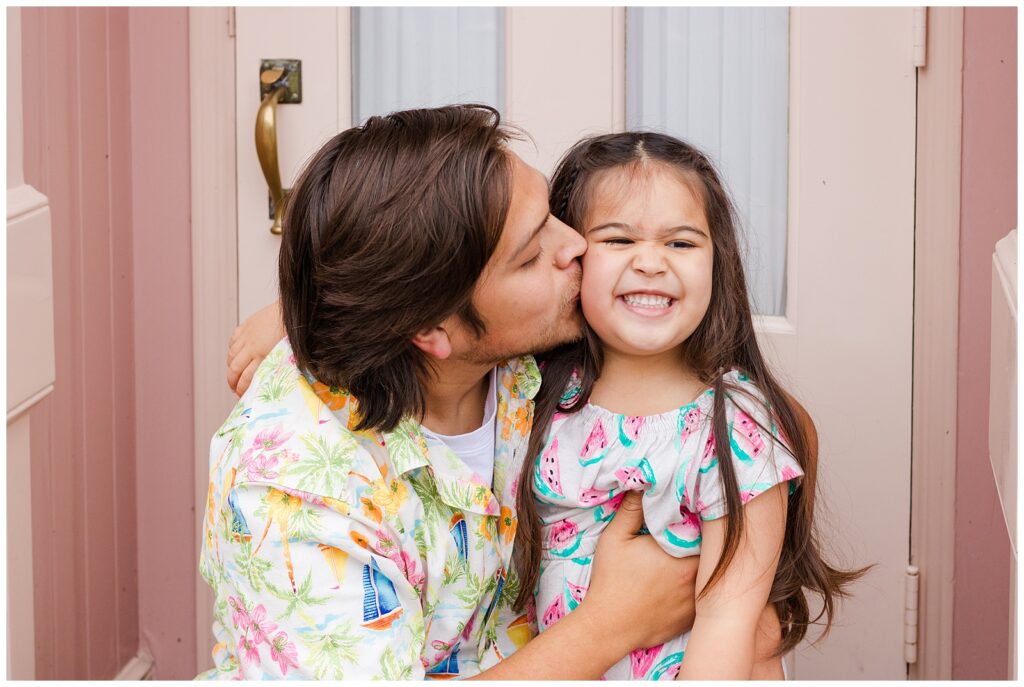 A father kisses his laughing daughter on the cheek in front of a pink storefront at Disneyland. The daughter is wearing a watermelon-patterned dress, and the father is dressed in a colorful floral shirt.