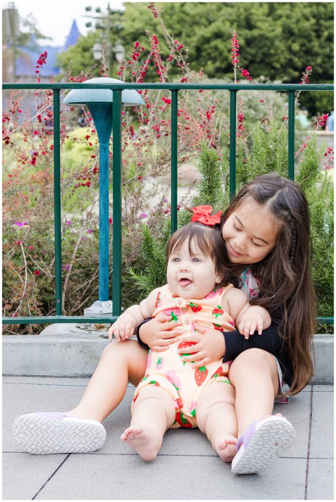 Two sisters sit on the ground in front of a fence with red flowers at Disneyland. The older sister hugs the younger baby sister, who is dressed in a strawberry-patterned romper with a red bow, and both are smiling.