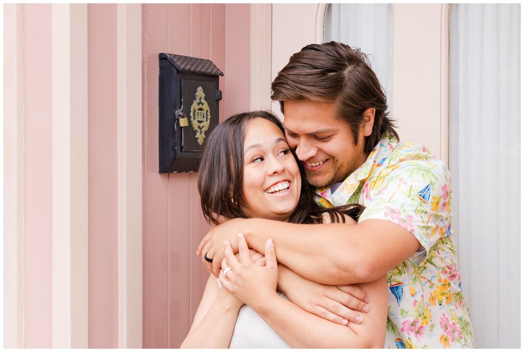 A couple embraces and smiles in front of a pink storefront at Disneyland. The man is wearing a colorful floral shirt, and the woman is dressed in a tank top and jeans. They look happy and in love.