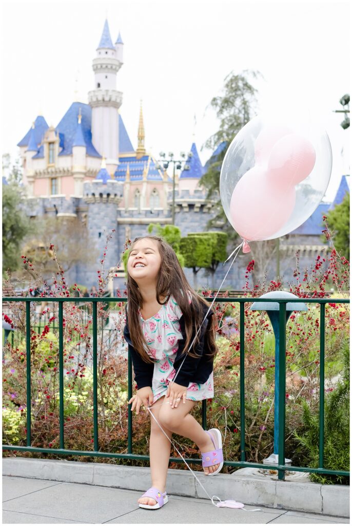 A young girl in a watermelon-patterned dress holds a Mickey Mouse balloon and laughs joyfully in front of Sleeping Beauty Castle at Disneyland. She is bending slightly forward, full of energy and happiness.