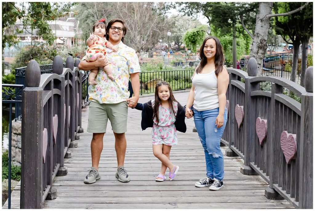 A family of four stands on a wooden bridge at Disneyland. The father holds the baby, dressed in a strawberry-patterned romper, while the mother and their older daughter, in a watermelon-patterned dress, hold hands. The family looks happy and united.