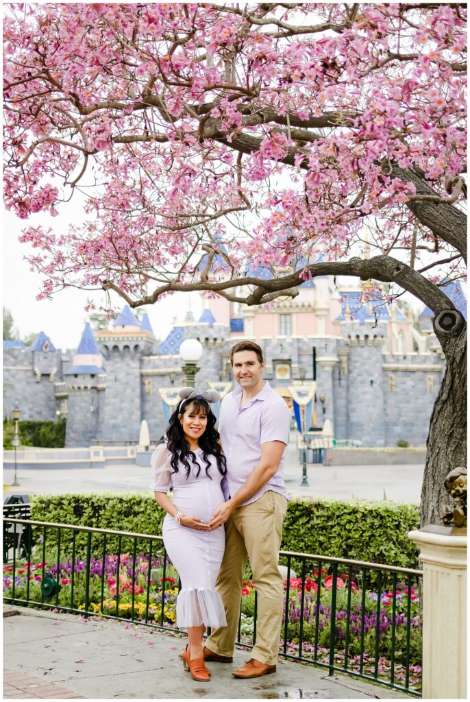 A couple stands together under a pink blossoming tree with Sleeping Beauty Castle in the background at Disneyland. The woman is pregnant, and they both smile warmly at the camera, with the man gently holding her from behind.