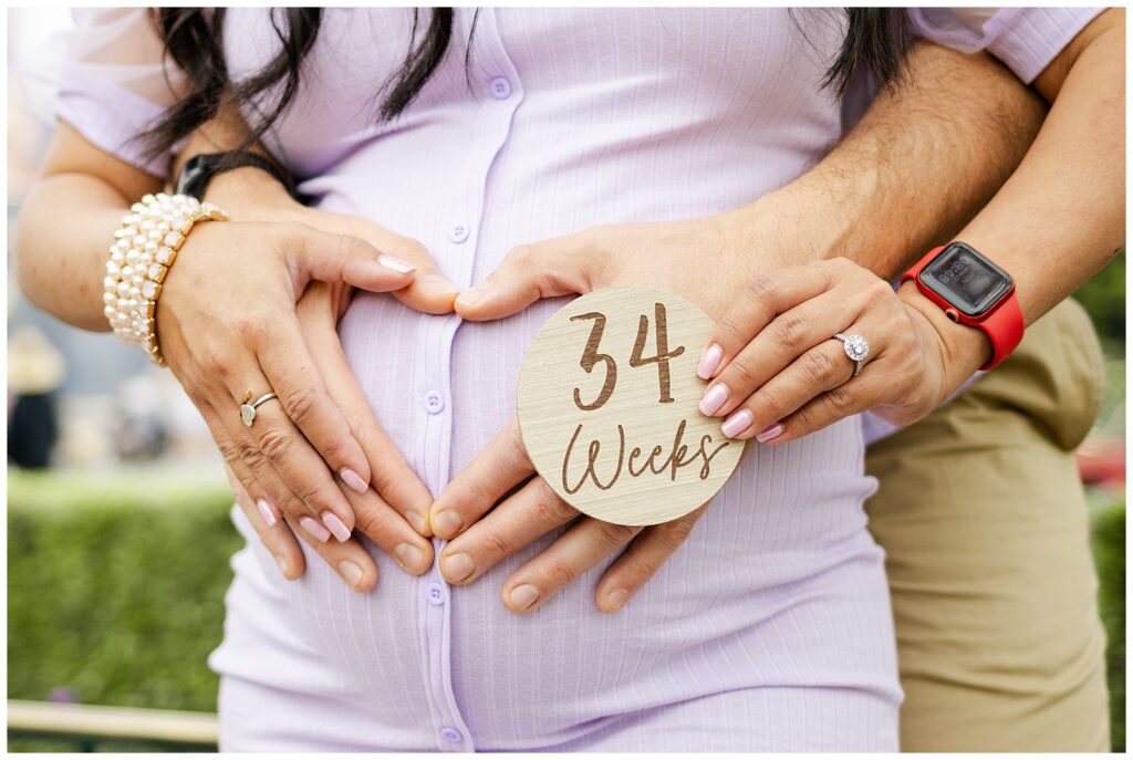 Close-up of a pregnant woman's belly, with her hands and her partner's hands forming a heart shape over her belly. They are holding a wooden sign that says "34 Weeks."