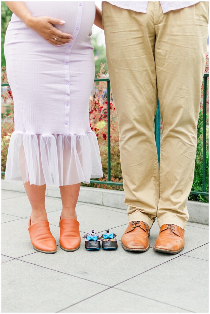 A pregnant couple stands together, showing only their lower bodies. The woman's pregnant belly is visible, and in front of them are tiny baby shoes placed on the ground, symbolizing the expected arrival of their baby.