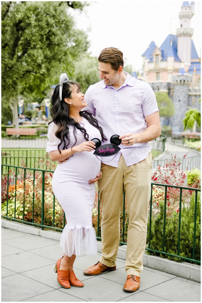 The couple stands close together in front of Sleeping Beauty Castle, holding a pair of Mickey Mouse ears with the words "Baby Hygge" embroidered in pink. They gaze lovingly at each other.