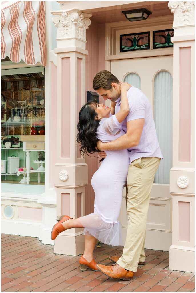 The couple shares a romantic moment, with the man dipping the woman and both smiling. They stand in front of a charming pink and white storefront, enhancing the Disney magic.