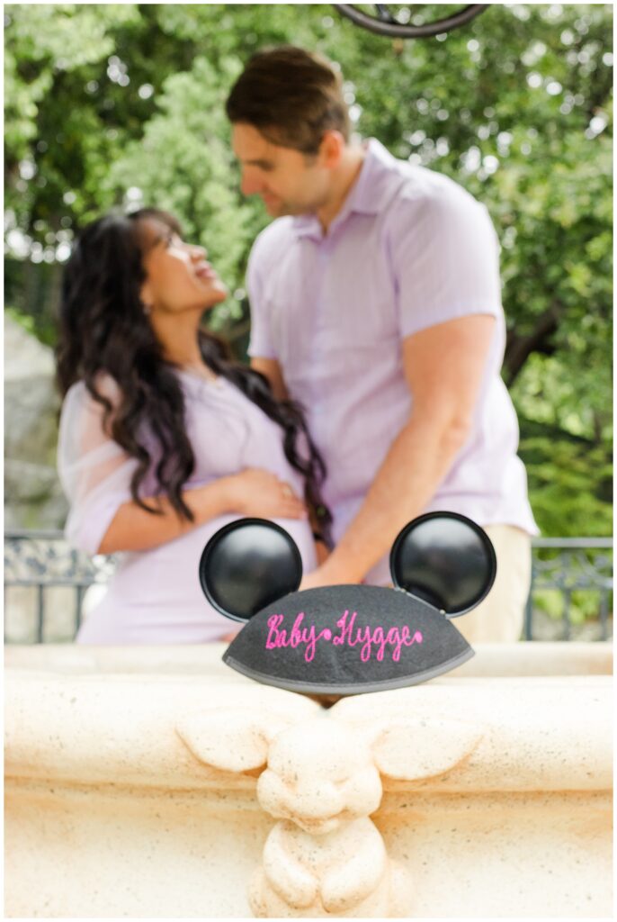 The couple stands together, blurred in the background, with Mickey Mouse ears in the foreground displaying "Baby Hygge." The photo focuses on the ears, symbolizing the upcoming addition to their family.