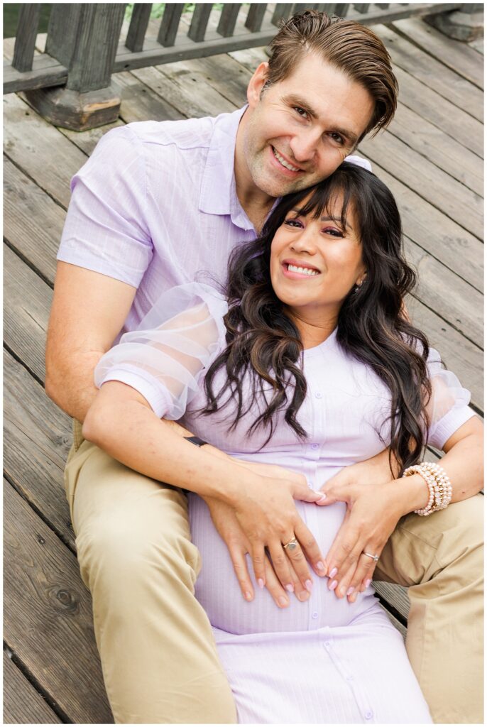 The couple sits together on a wooden bridge, with the woman leaning back against the man. They form a heart shape with their hands over her belly, both smiling contentedly at the camera.