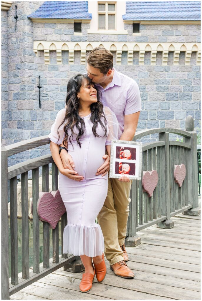 The couple stands on a wooden bridge in Disneyland, with the man kissing the woman's forehead. She holds her belly and a picture frame with ultrasound images, both smiling and looking happy.