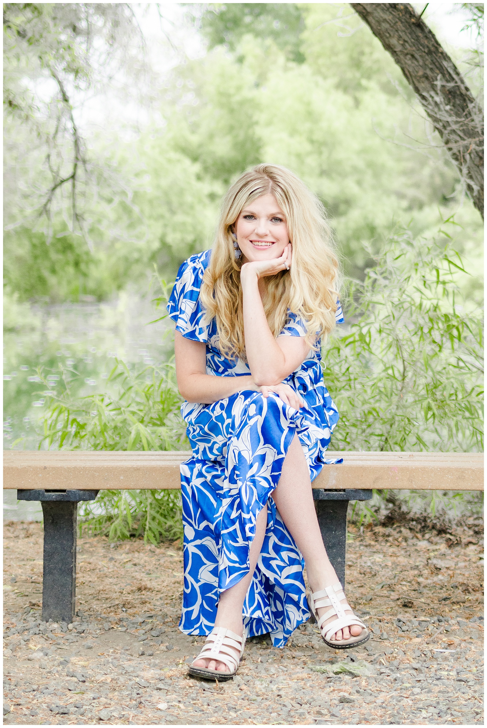 A blonde woman wearing a blue and white floral dress is sitting on a bench in a natural, wooded area. She is smiling and resting her chin on her hand, looking at the camera. She is wearing white sandals.