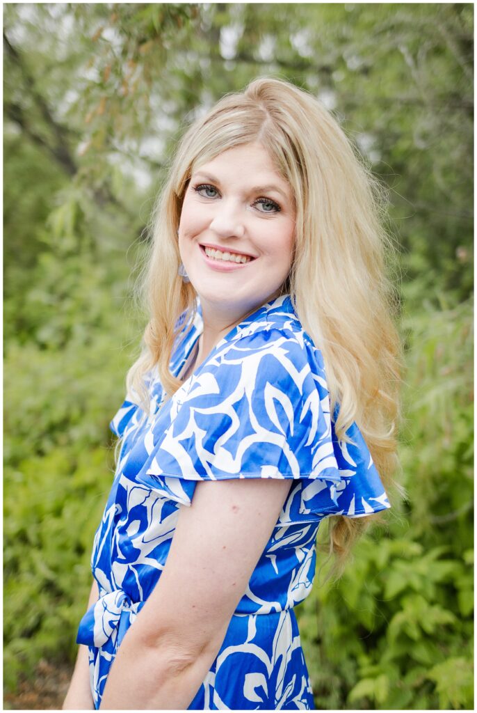 A blonde woman wearing a blue and white floral dress is standing outdoors in a green, leafy area. She is smiling at the camera and her hair is styled in loose waves.