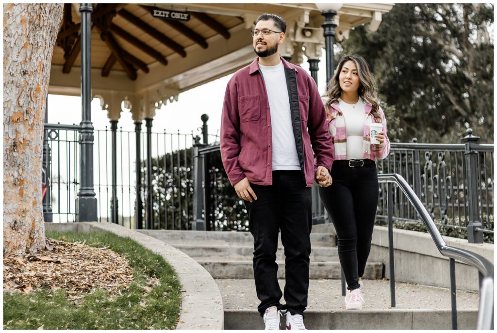 A couple holds hands as they walk down steps in a park, with the man looking ahead and the woman glancing to the side. They are dressed in casual, coordinated outfits, and the park's gazebo and trees provide a picturesque backdrop.