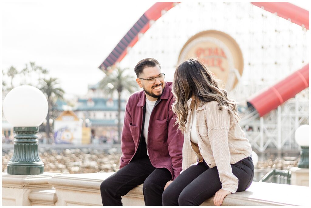 A couple sits on a ledge with a theme park ride in the background. They both smile warmly at each other, enjoying the sunny day and the excitement of the park.