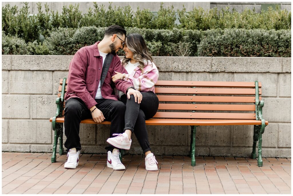 A couple sits close together on a bench, sharing a tender moment. The man leans in towards the woman, who looks at him lovingly, with a row of green bushes behind them.