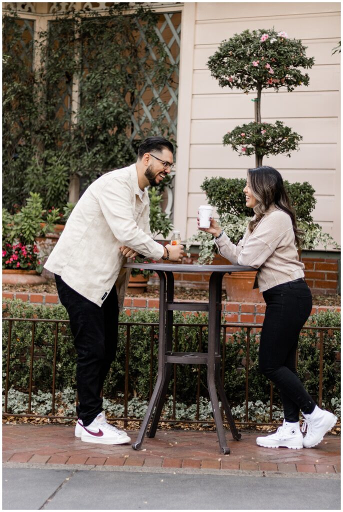 couple stands at a tall table, smiling and enjoying drinks together. The man leans on the table, holding a bottle, while the woman stands across from him, holding a cup. They are surrounded by greenery and a quaint, decorative background.