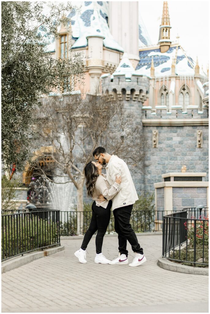 A couple shares a kiss in front of a castle, surrounded by lush greenery. The man embraces the woman as they lean in for a kiss, with the castle's beautiful architecture visible behind them.