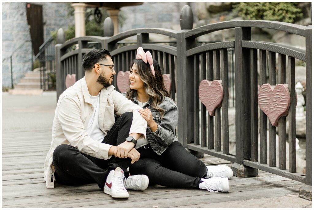 A couple sits on a wooden bridge, surrounded by heart-shaped decorations on the railing. They look at each other with smiles, enjoying a peaceful, intimate moment.