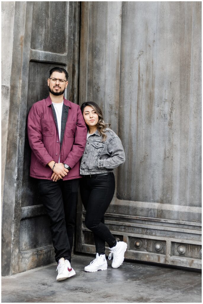 A couple stands together, leaning against a large metallic door. The man wears a burgundy jacket and glasses, while the woman stands beside him in a gray denim jacket, both looking relaxed and stylish.